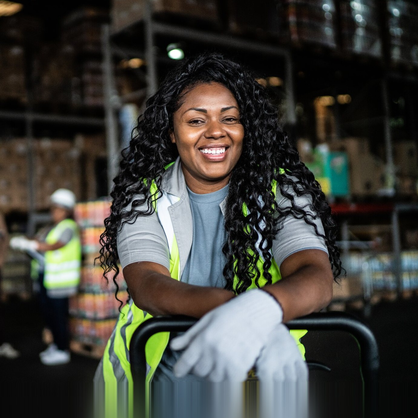 Smiling warehouse worker in a hi-vis vest leaning on a trolley, colleagues behind